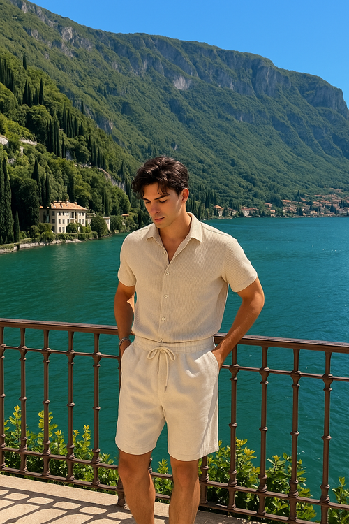 Ein Mann in einem beigefarbenen, gerippten Strick-Set aus Kurzarmhemd und Shorts mit Kordelzug, steht auf einem Balkon mit Blick auf einen malerischen See und grüne Berge.